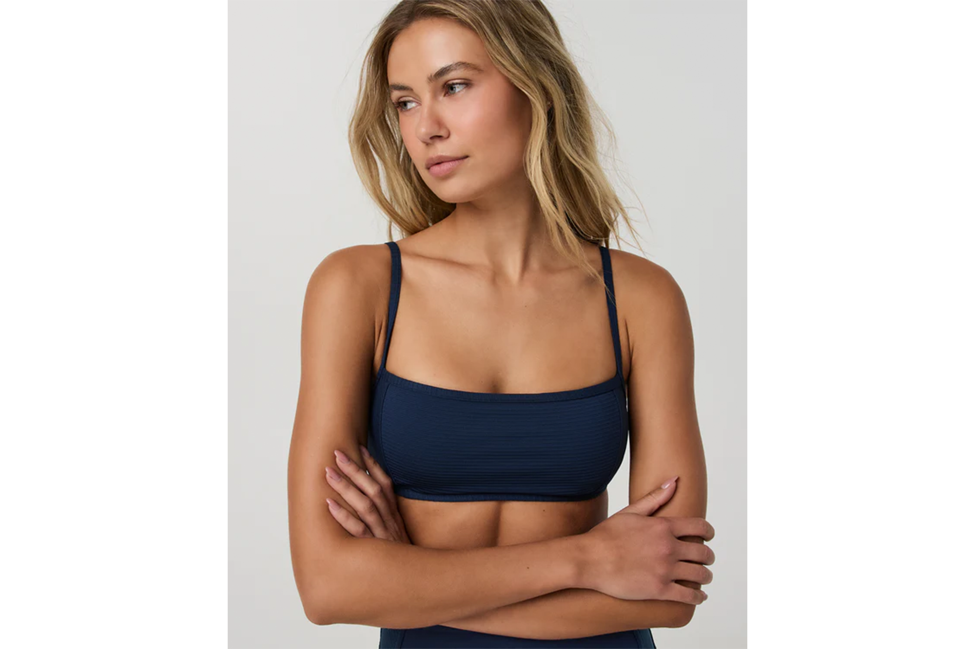 A woman wearing a navy blue ribbed athletic bra with thin straps, looking to the side with arms crossed.