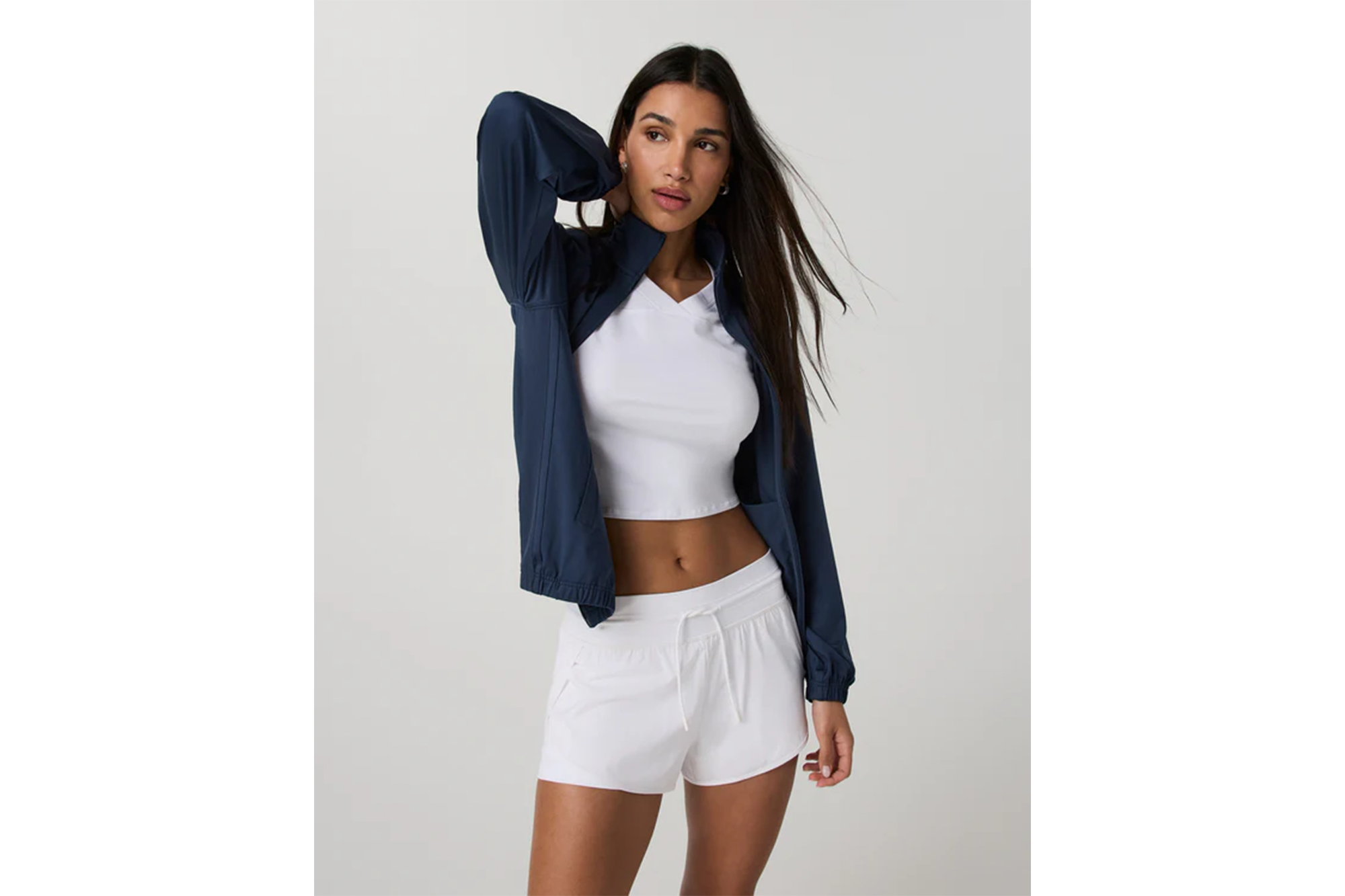 A model poses in a navy blue zip-up jacket, white crop top, and white shorts.