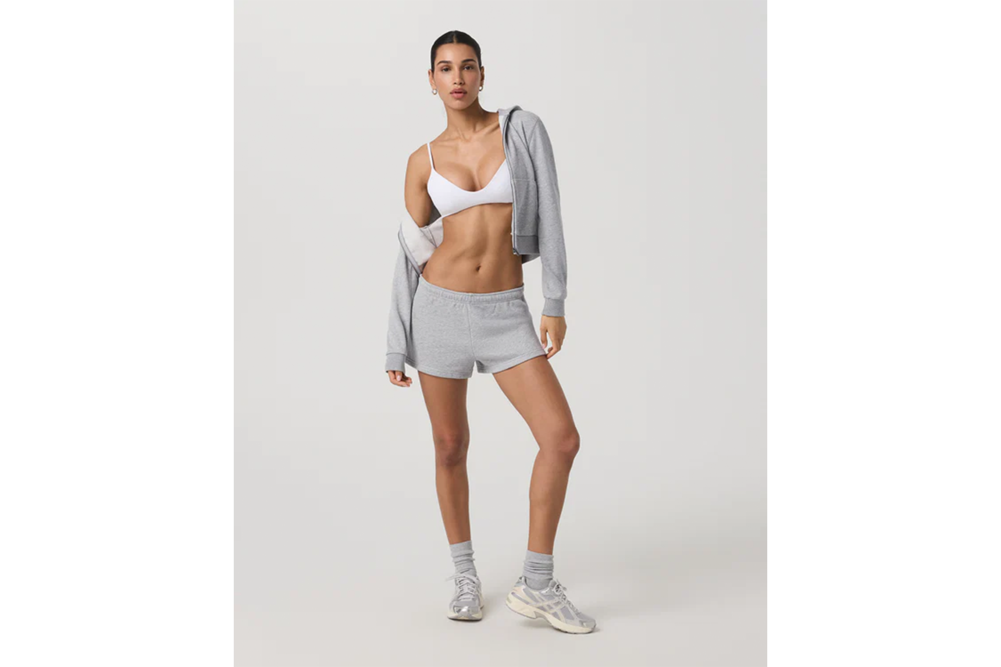 A woman wears a white sports bra, gray shorts, a gray zip-up hoodie draped over her shoulders, and sneakers.