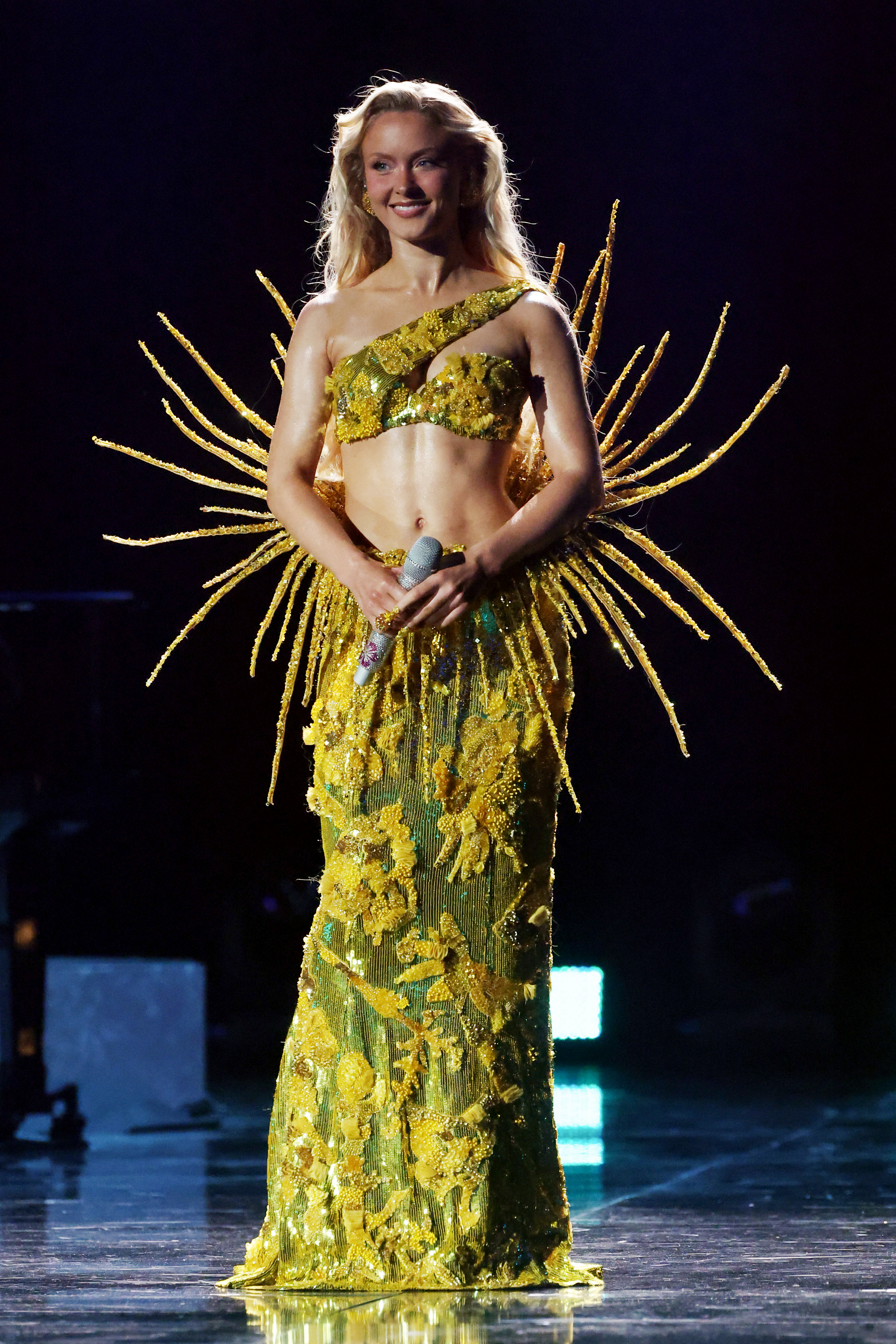 Zara Larsson performing onstage at the 68th GRAMMY Awards Premiere Ceremony, wearing a shimmery golden two-piece outfit with spiky protrusions.