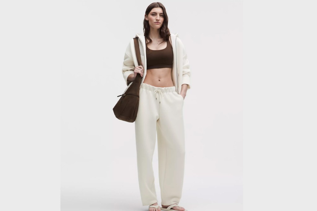 A woman wearing a brown sports bra, a cream-colored zip-up hoodie, cream sweatpants, and a brown sling bag.