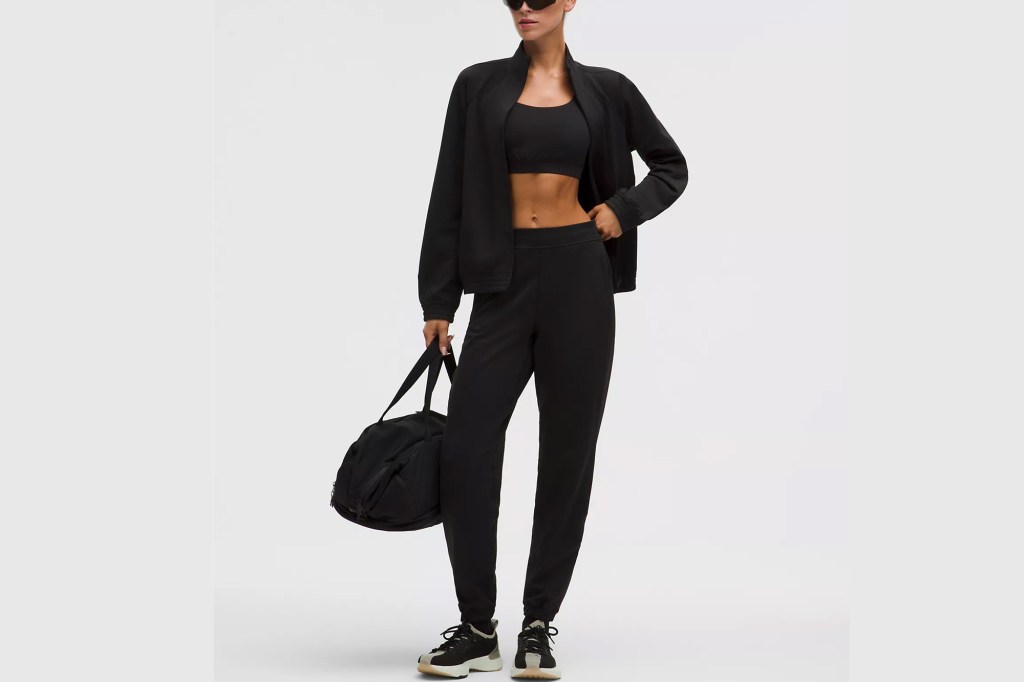 A model wearing a black full-zip bomber jacket, black sports bra, black athletic pants, and black duffel bag.