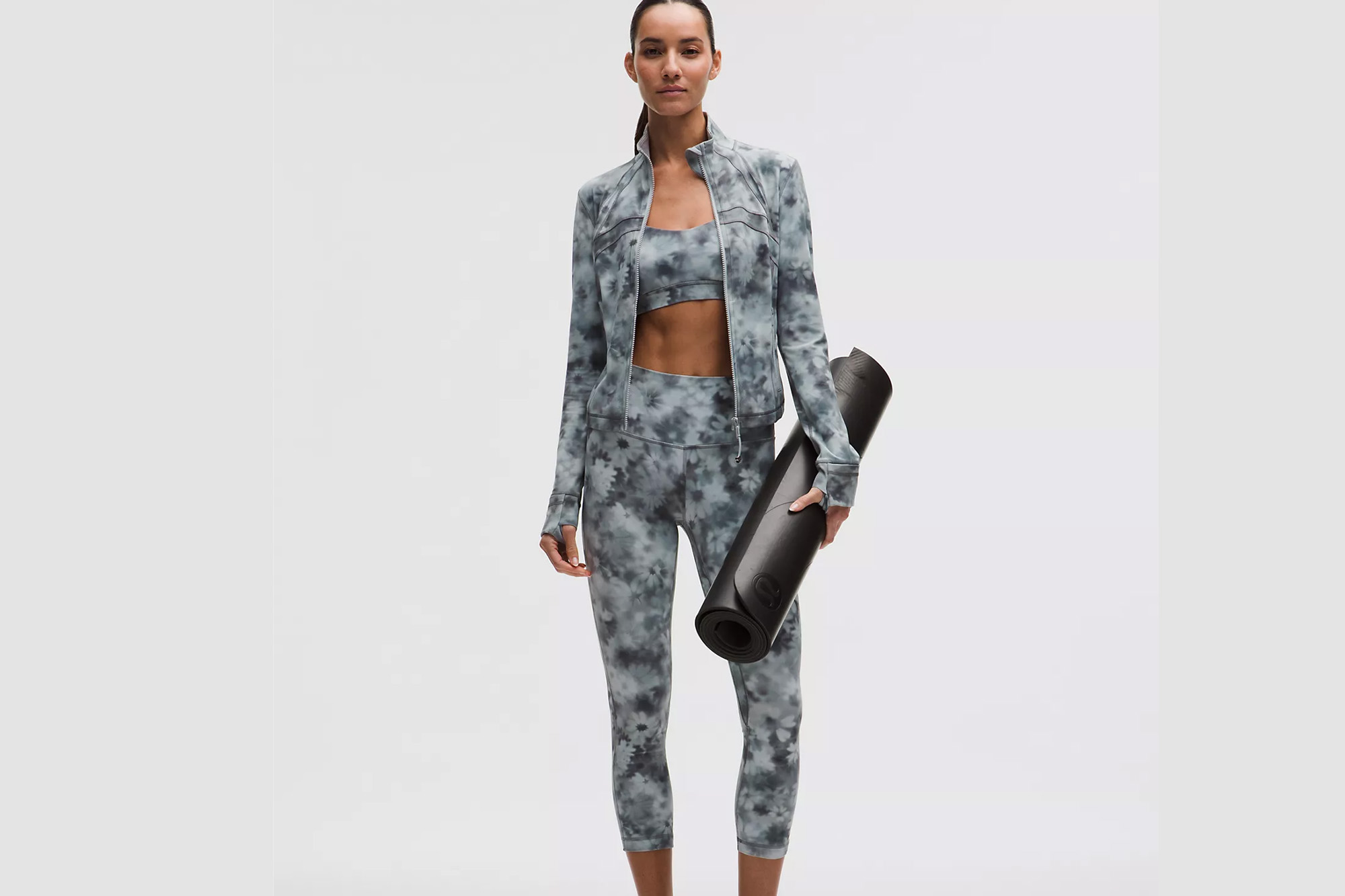 A woman in a gray and white tie-dye athletic set holds a black yoga mat.