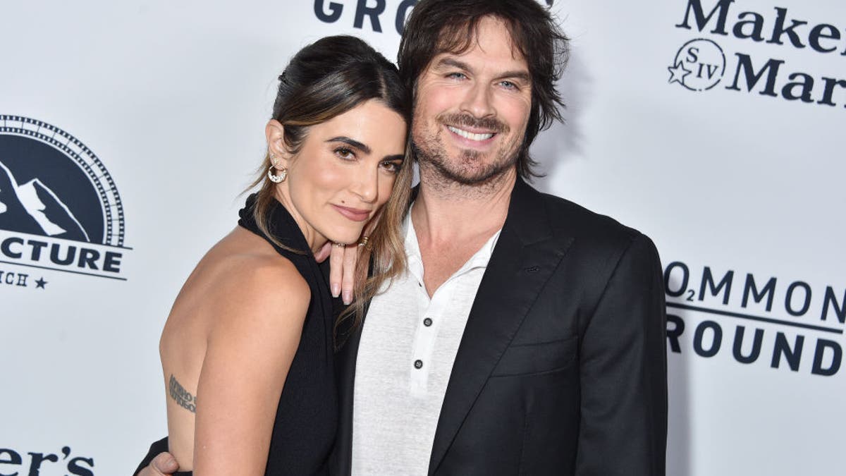 Nikki Reed in black halter dress leans into Ian Somerhalder in black jacket on carpet