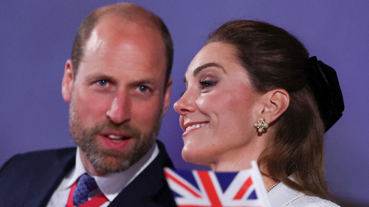 Prince William speaking to Catherine, Princess of Wales, with a Union Jack flag in front of her