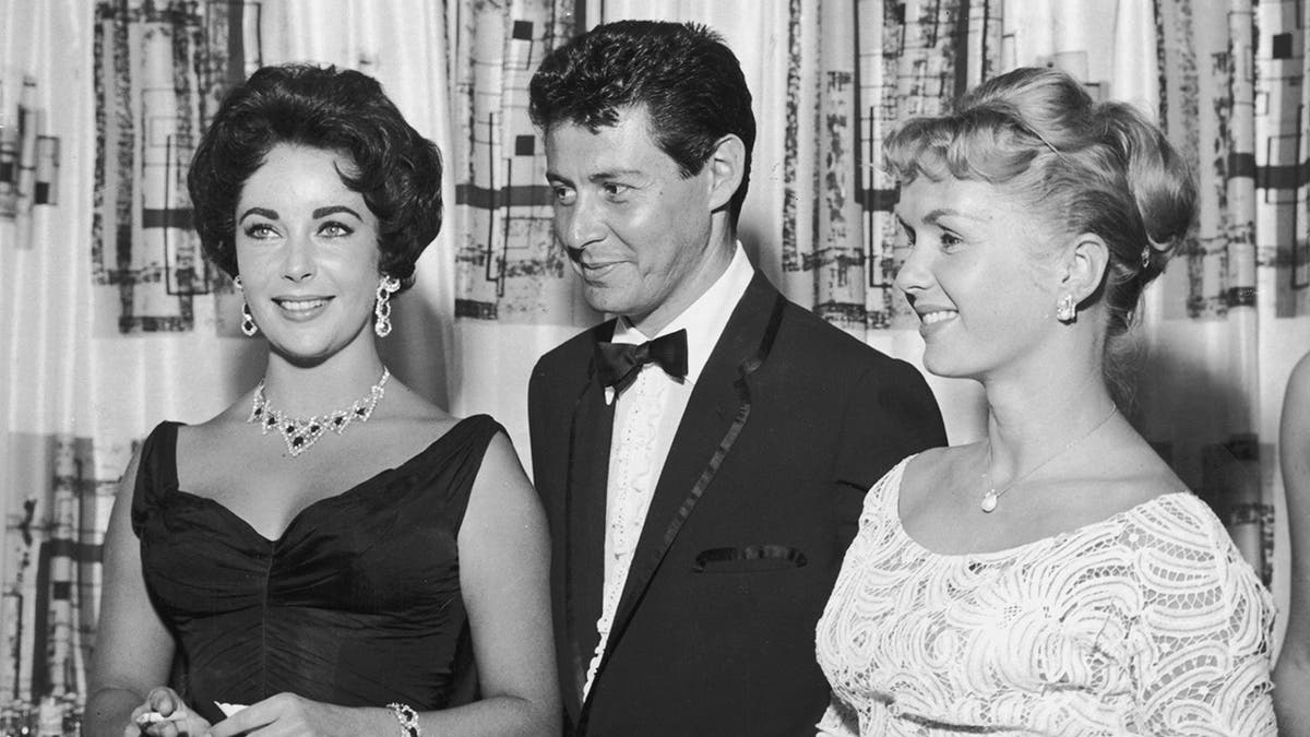 Eddie Fisher standing with arm around Debbie Reynolds while looking at Elizabeth Taylor smoking a cigarette