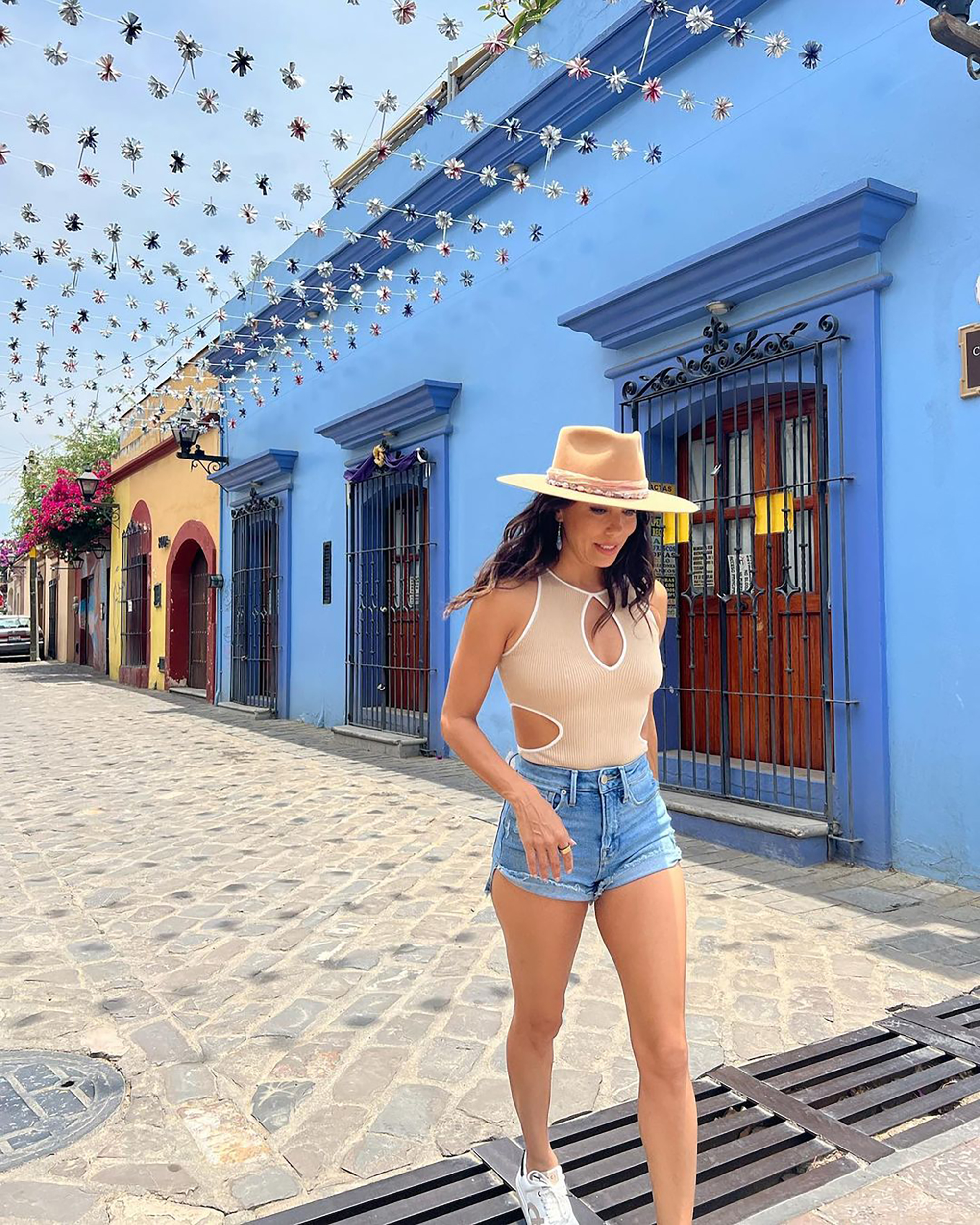 Eva Longoria in a beige cutout top, denim shorts, and a hat walks on a cobbled street lined with colorful buildings under sky decorations.