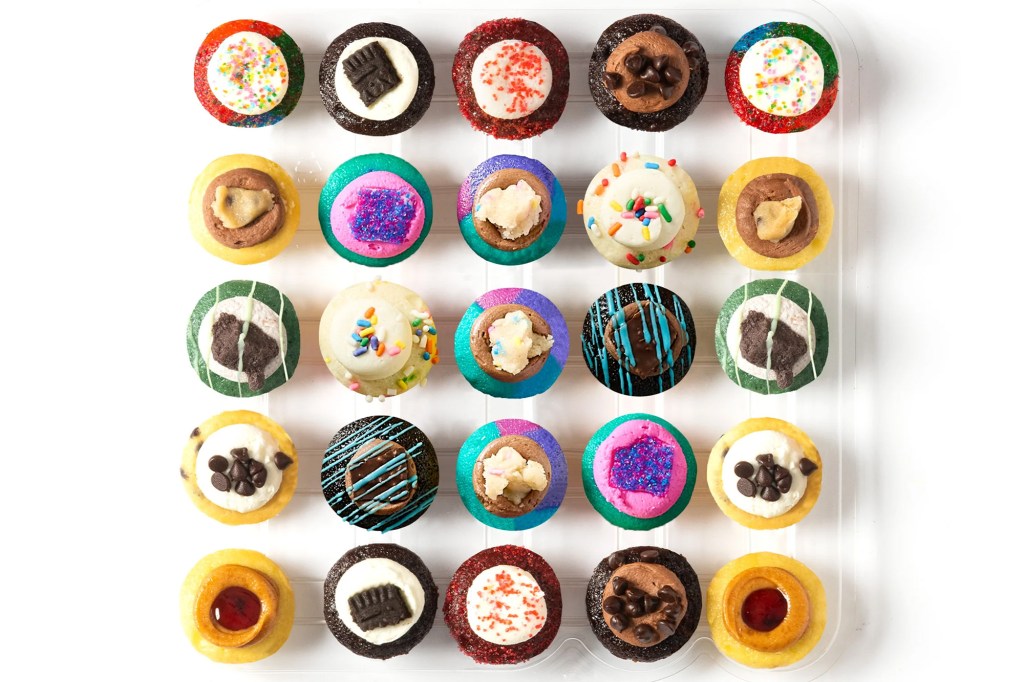 An overhead shot of 25 cupcakes with various frostings and toppings arranged in rows within a clear plastic container.