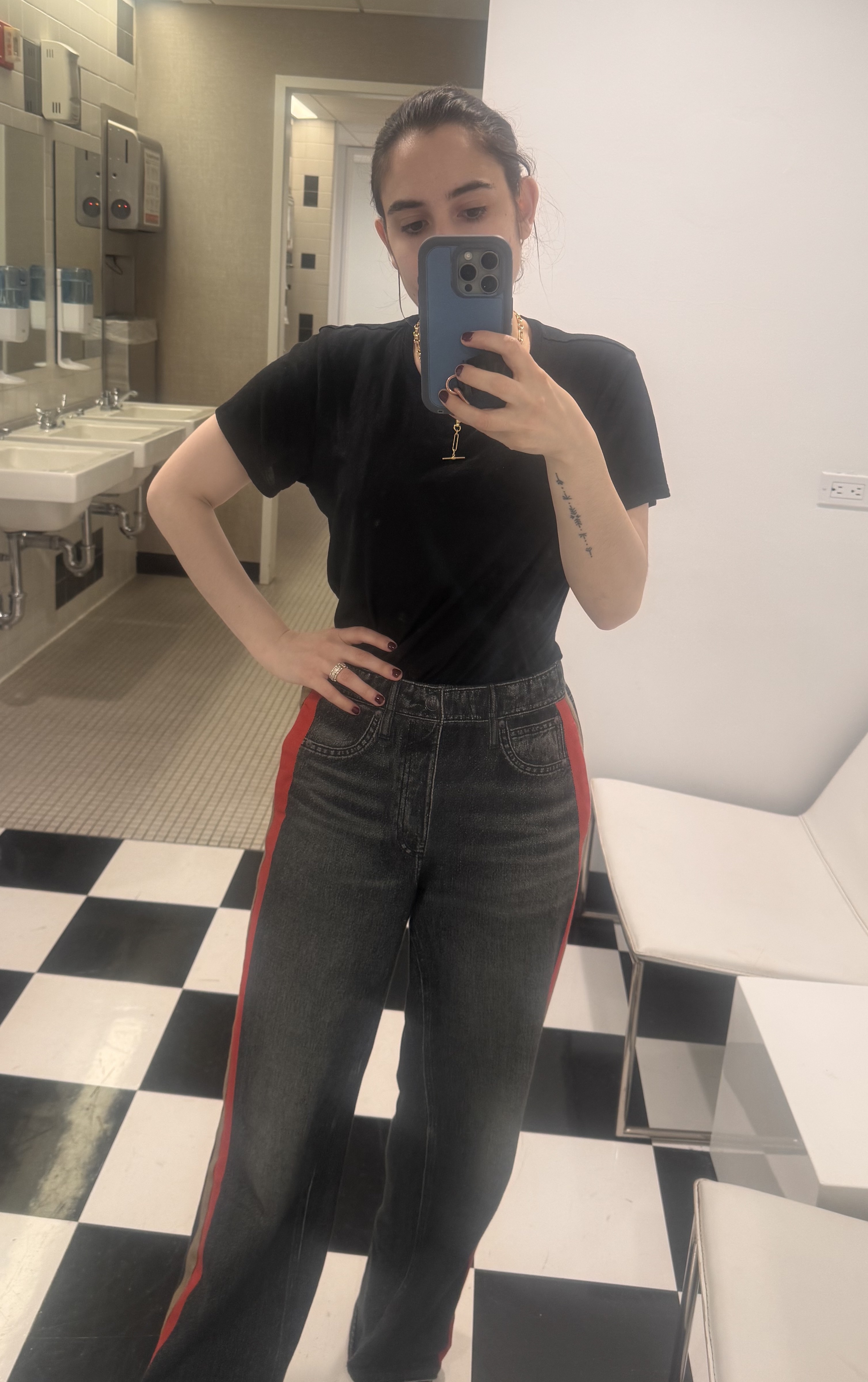 Woman in a black t-shirt and dark wash jeans with red stripes taking a mirror selfie in a restroom.