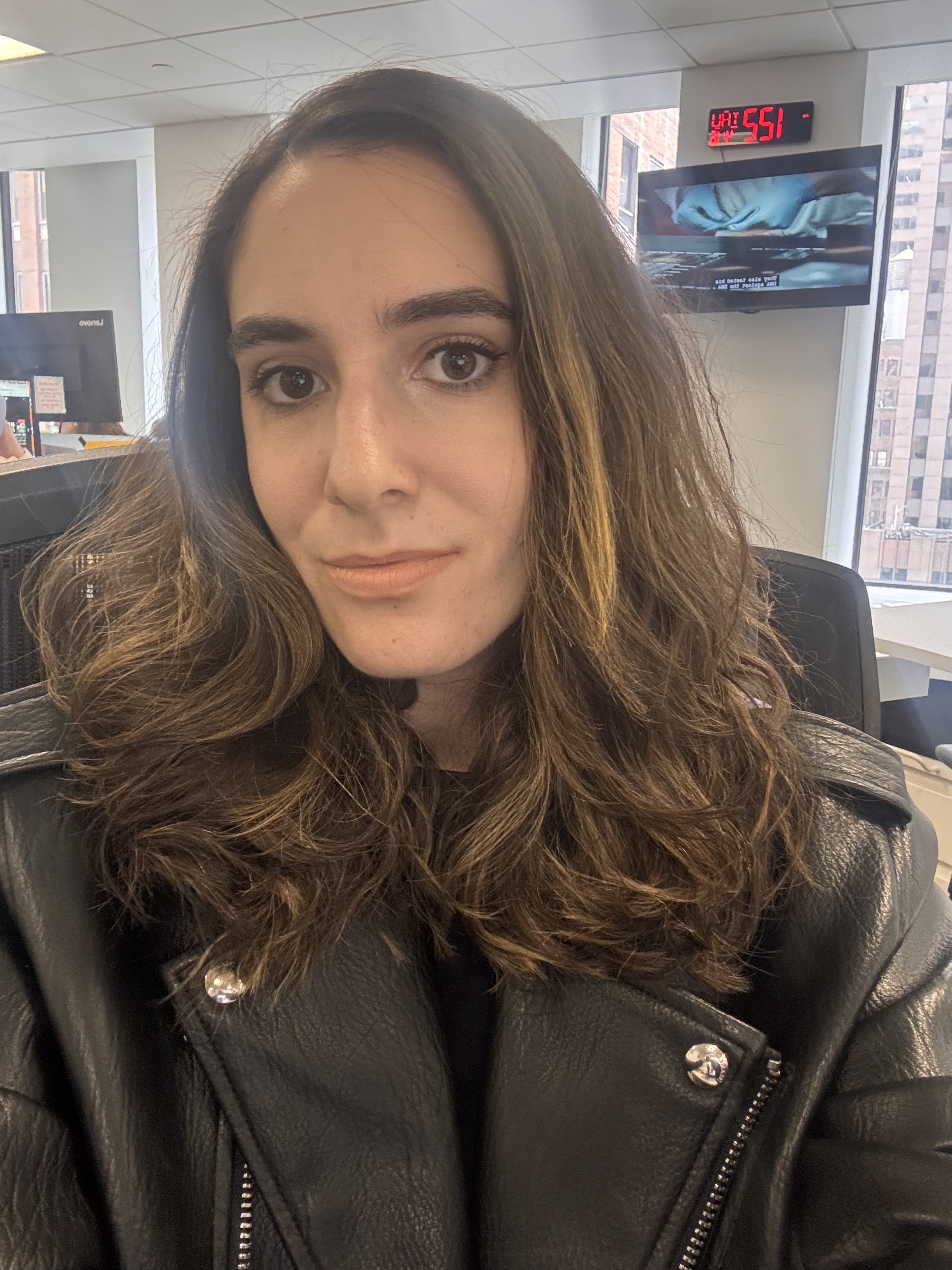 A woman with long wavy brown hair and wearing a black leather jacket takes a selfie in an office.