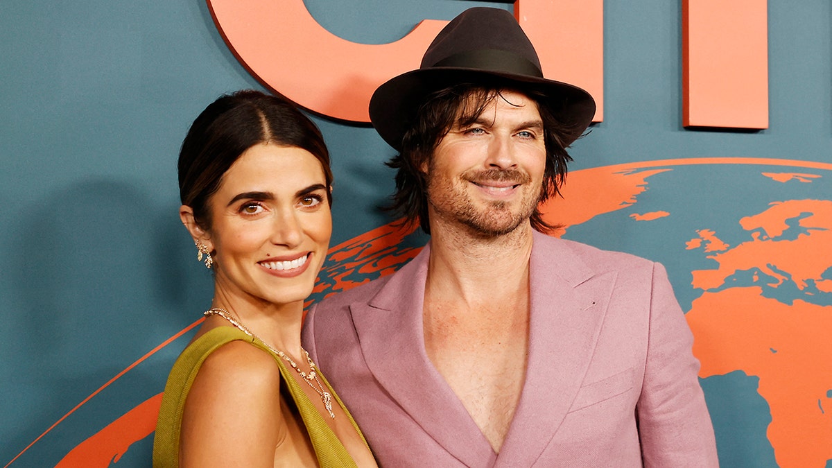 Nikki Reed smiling in a green dress with Ian Somerhalder in a light pink blazer and brown hat at an awards gala