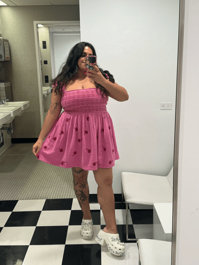 A woman with dark hair wearing a pink dress and embellished white platform Crocs takes a selfie in a mirror.