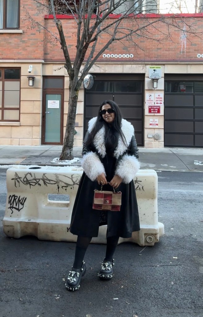 Woman wearing a black coat with white fur trim, dark sunglasses, and studded platform shoes, holding a checkerboard patterned handbag.