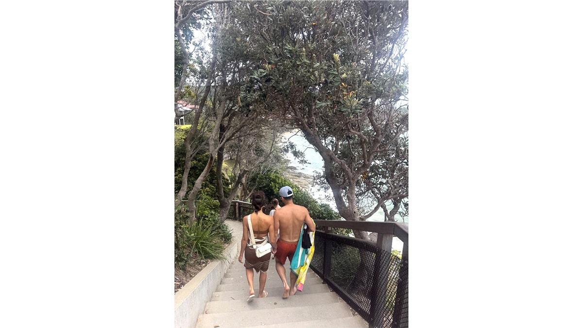 Jessica Alba and Danny Ramirez holding hands walking down a tree-lined staircase toward the beach.