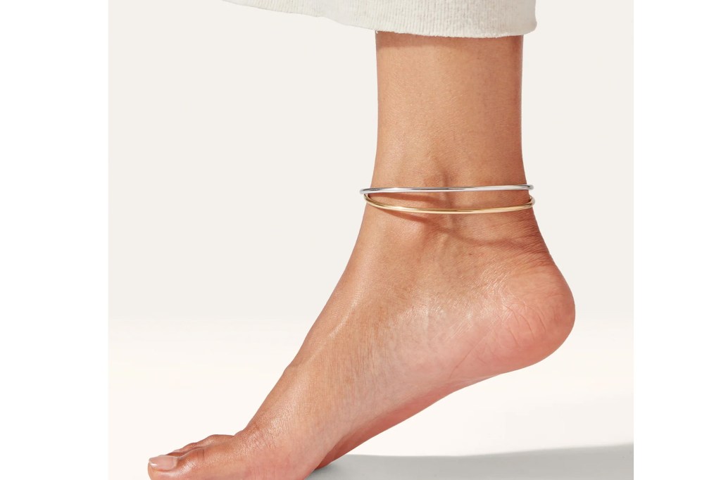 A woman's ankle with a silver and a gold-colored ankle bracelet stacked on it.