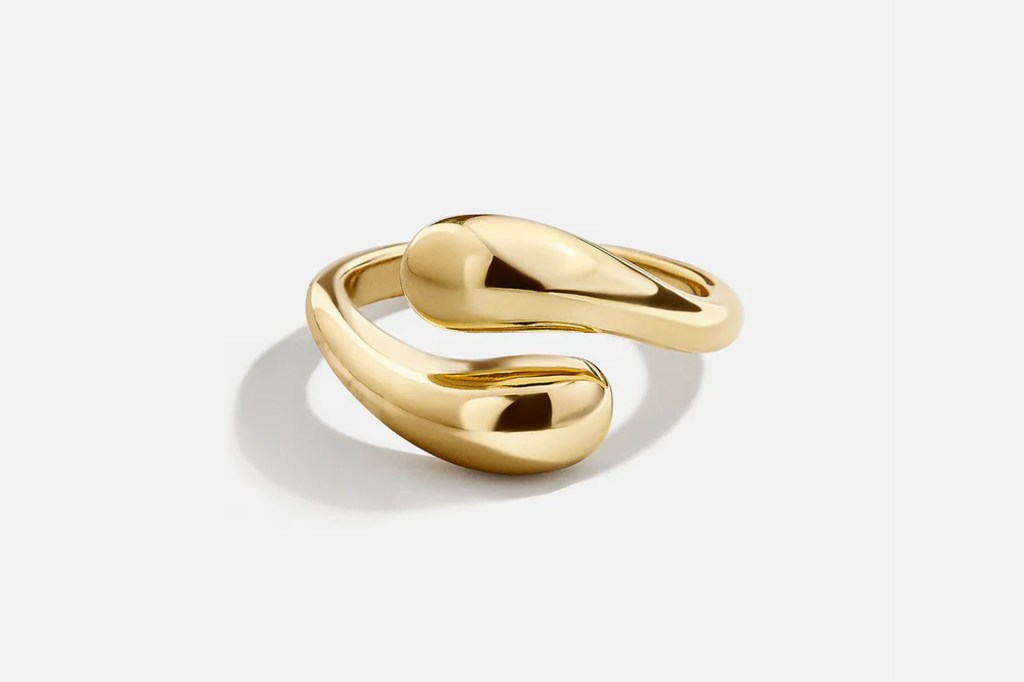 A gold ring with a thick, sculptural, open-ended design, featuring two teardrop shapes at the ends, on a white background.