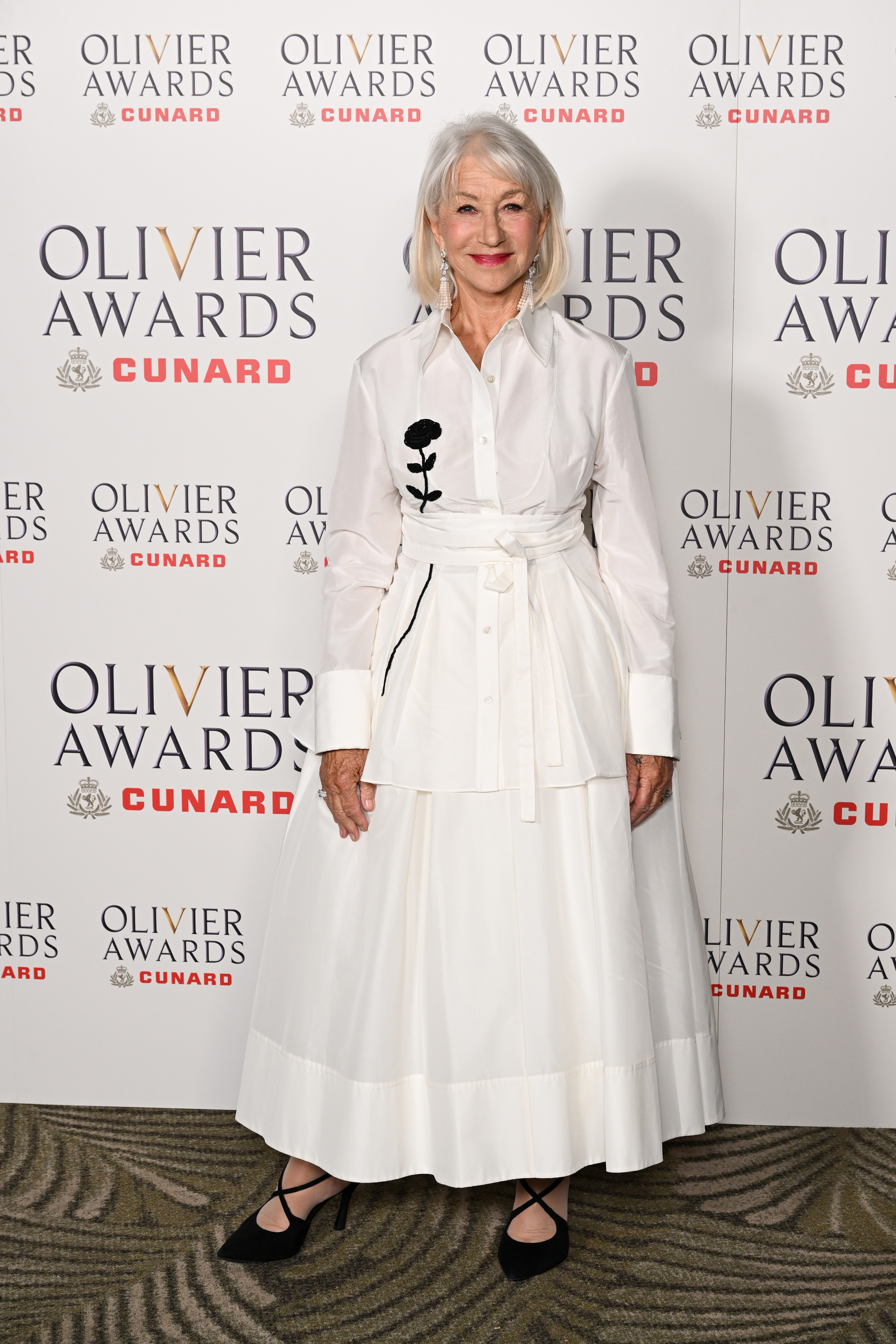 Dame Helen Mirren poses in a white long-sleeved dress with a black embroidered rose, at The Olivier Awards with Cunard.