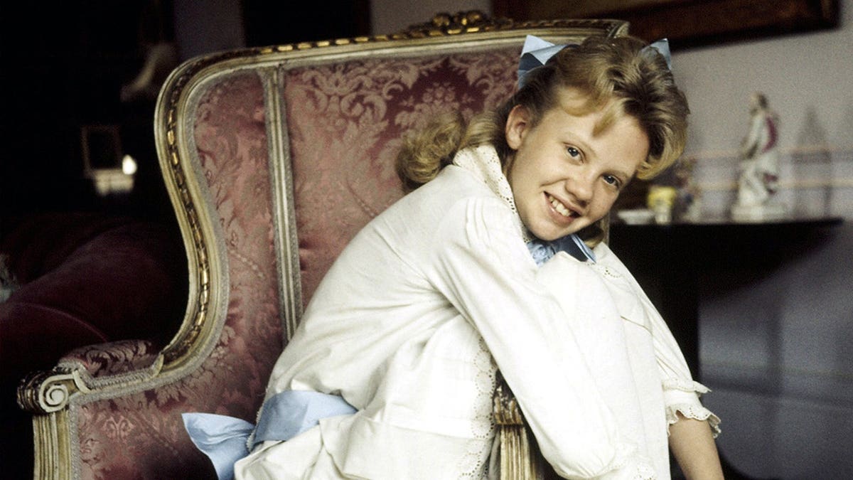Hayley Mills sitting in a chair wearing a white dress with a blue sash belt.