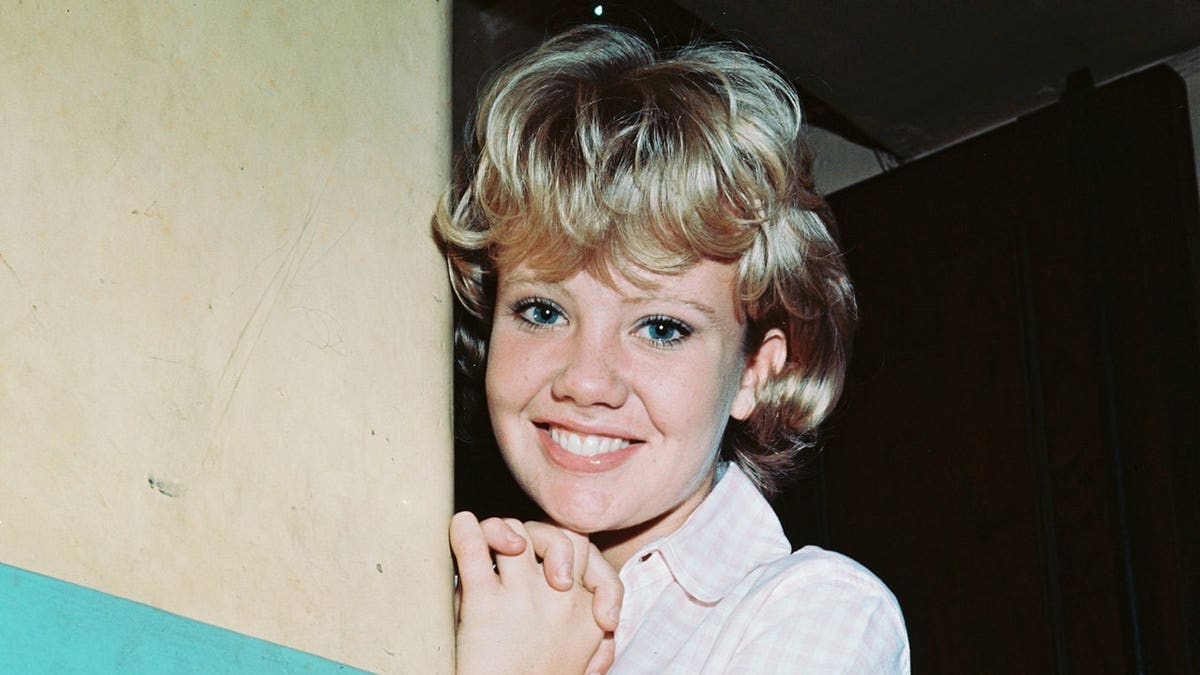 Hayley Mills smiling and posing against a blue and white wall wearing a white dress with a red belt