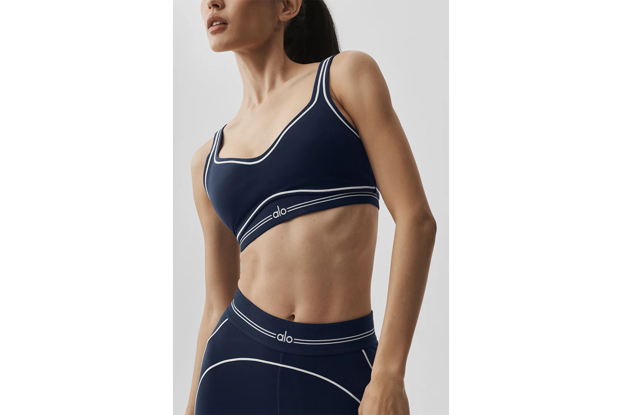 A woman wears a navy blue sports bra and matching leggings with white trim and "alo" branding.