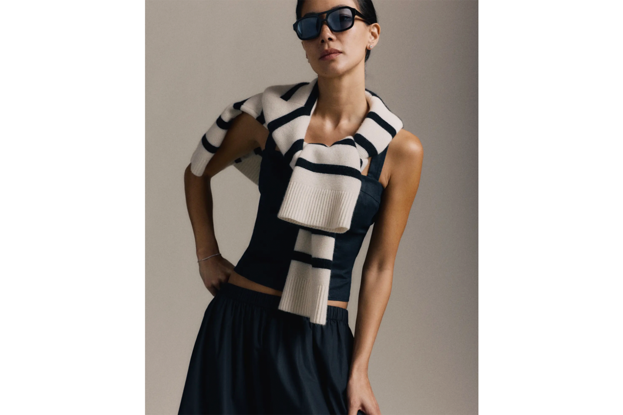 A model in a black top, dark blue skirt, and sunglasses, with a cream and black striped sweater draped over her shoulders.