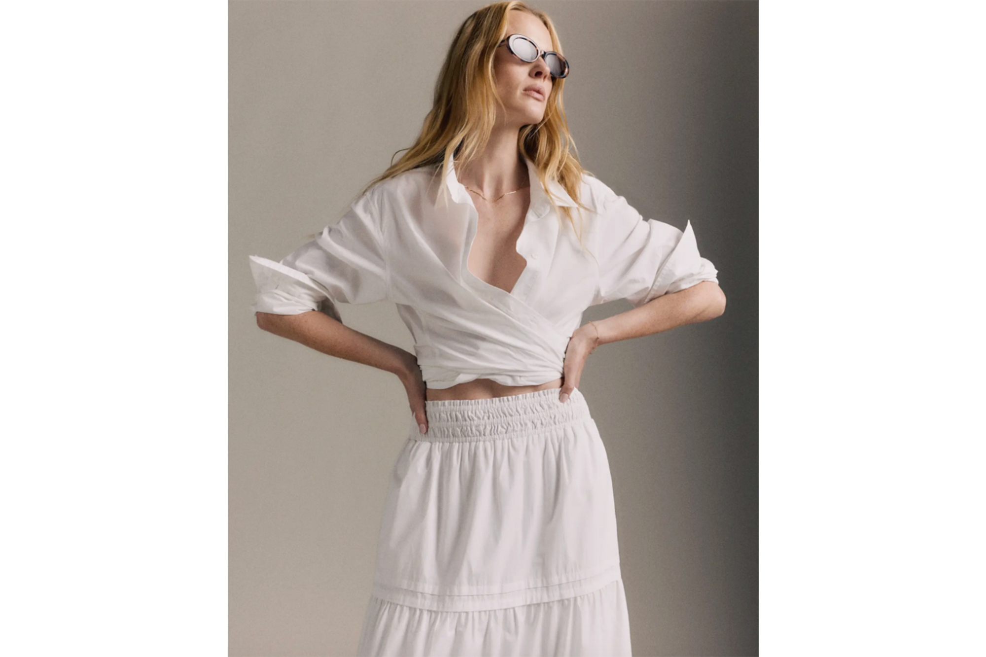 A woman wearing an unbuttoned white shirt tied around her waist and a white tiered skirt, looking up while wearing sunglasses.