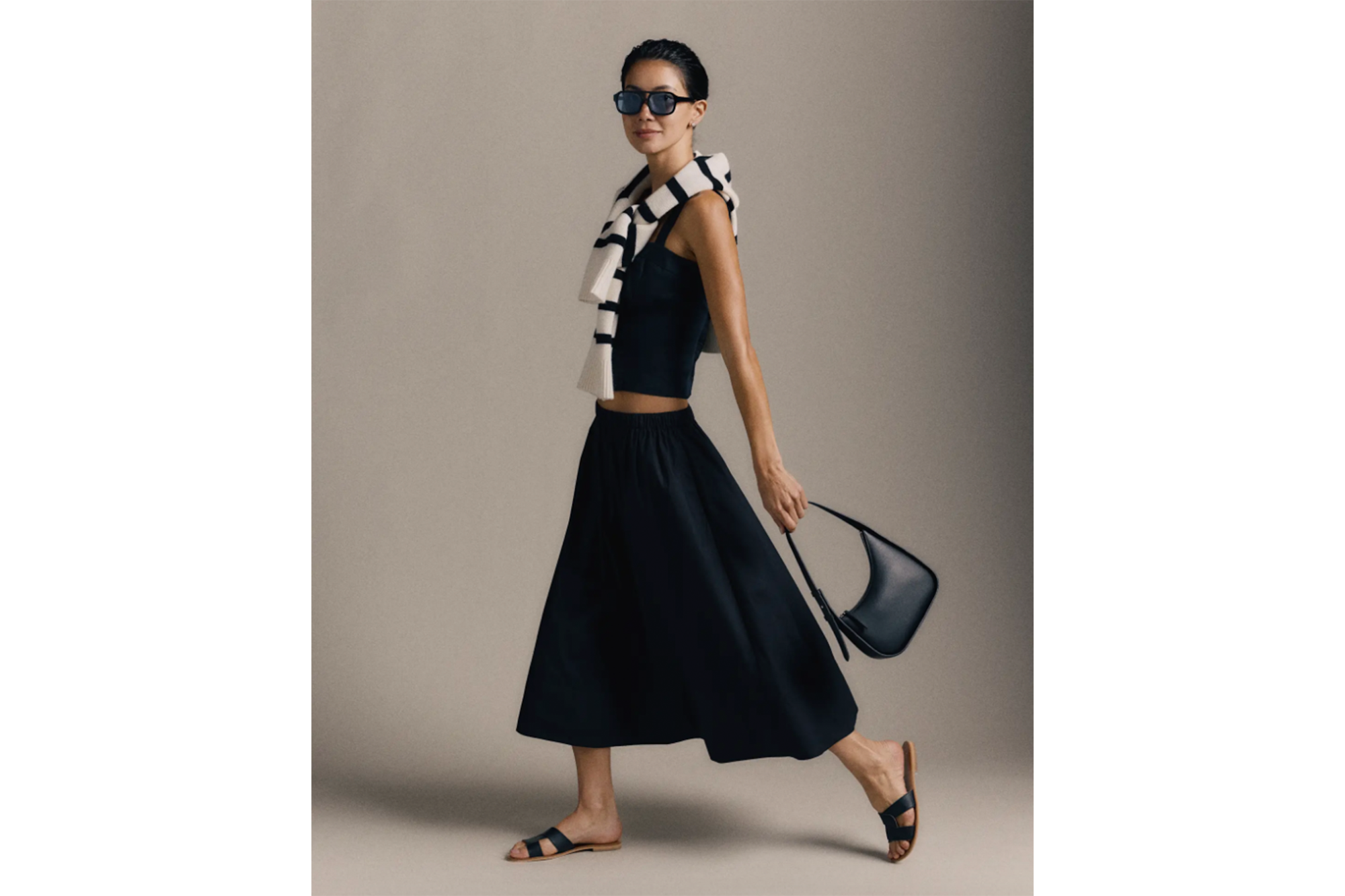 A woman wearing a black crop top and flowing black midi skirt, black flat sandals, sunglasses, and carrying a black purse, with a striped sweater draped over her shoulders.