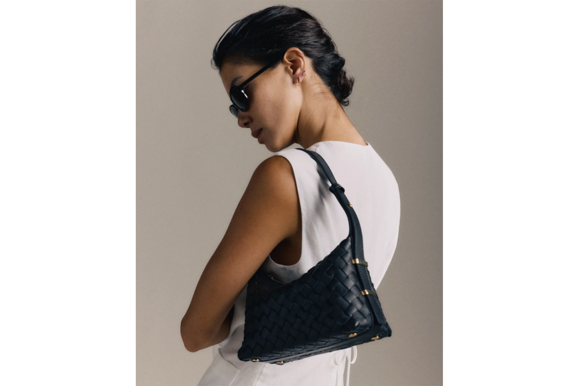 Woman wearing sunglasses and a white top, holding a black woven leather shoulder bag.