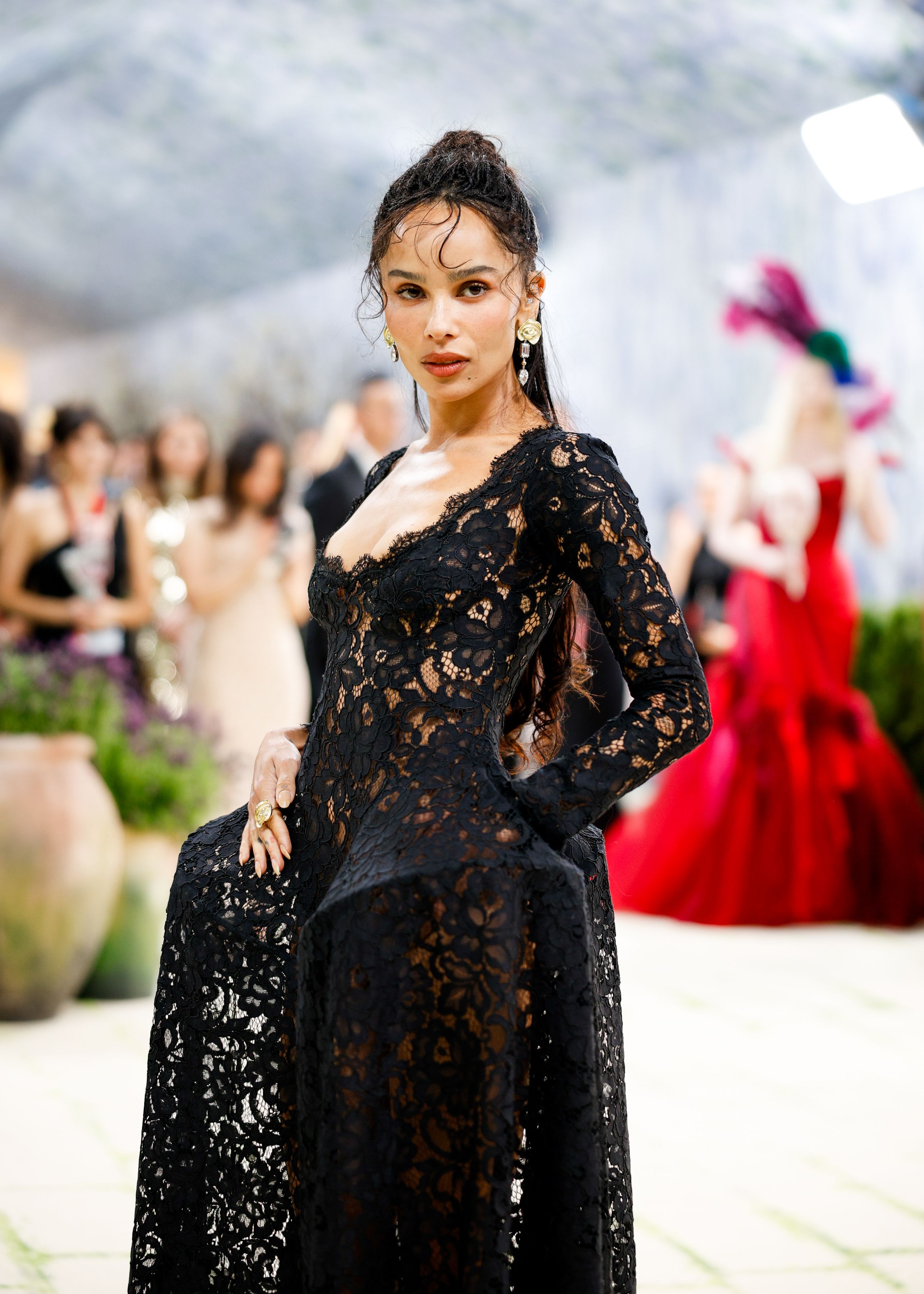 Zoë Kravitz posing in a black lace dress with a ruffled skirt, gold earrings, and her hair slicked back.