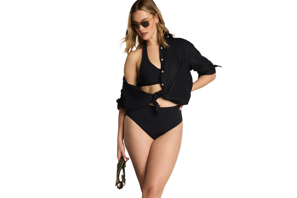 A woman wearing a black halter bikini top and high-waisted bottoms, with a black collared shirt tied at her midriff, and sunglasses.