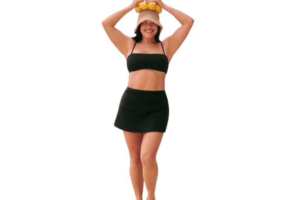A woman in a black swimsuit and black swim skirt balancing a woven straw hat filled with lemons on her head.