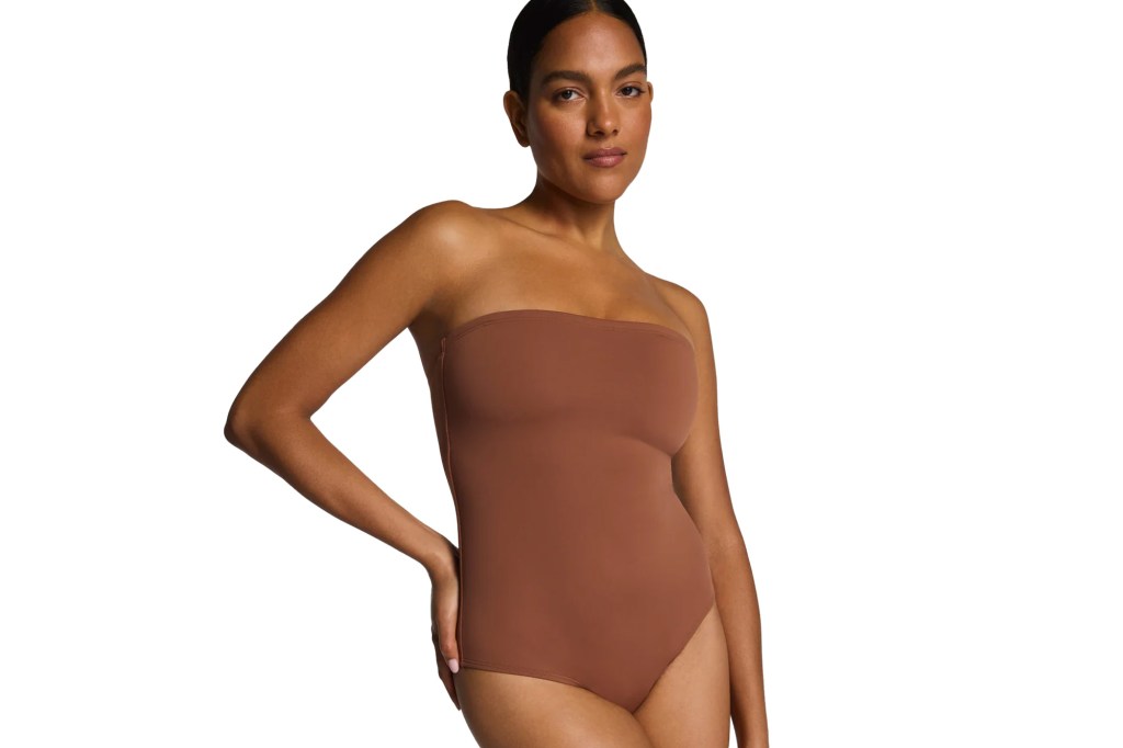 A woman with dark skin in a strapless brown bodysuit looks directly at the viewer, with her left hand on her hip.