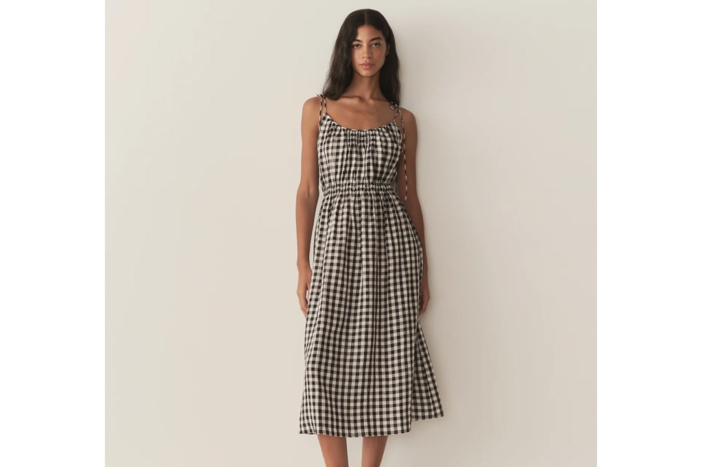 A woman with long dark hair stands facing the camera, wearing a black and white gingham midi dress with spaghetti straps and a shirred waist.