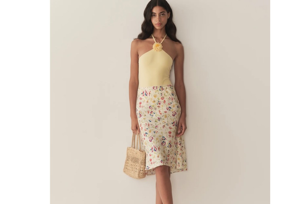 A woman models a yellow halter top with a flower appliqué and a white floral skirt, holding a woven straw bag.