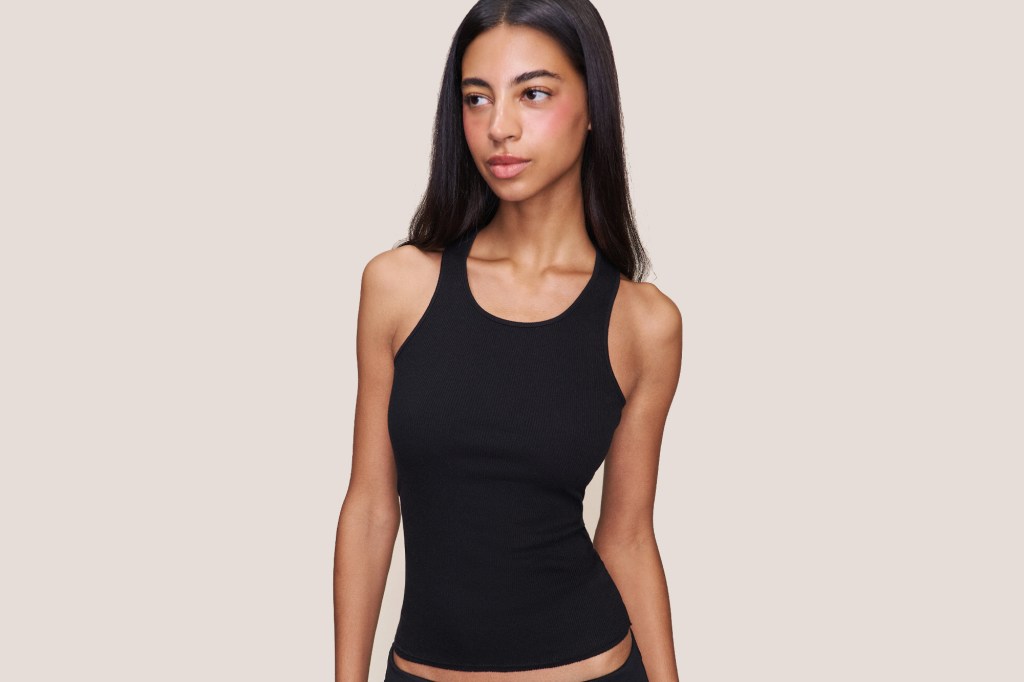 A model with long dark hair, wearing a black ribbed tank top, looks to the right.