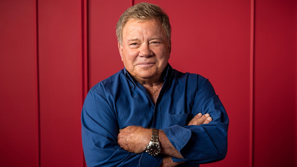 William Shatner posing with arms crossed in a blue shirt at a portrait studio.