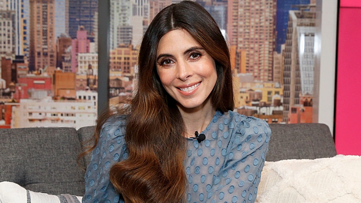 Jamie-Lynn Sigler sitting on a couch wearing a light blue lace dress and smiling